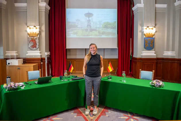 Person posing in front of two green tables in an old aula