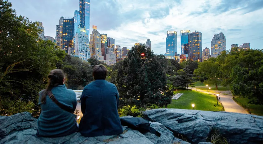 A man and a woman are phorographed from behind, sitting on a big rock in a city park at dusk, gazing up at the skyscrapers  