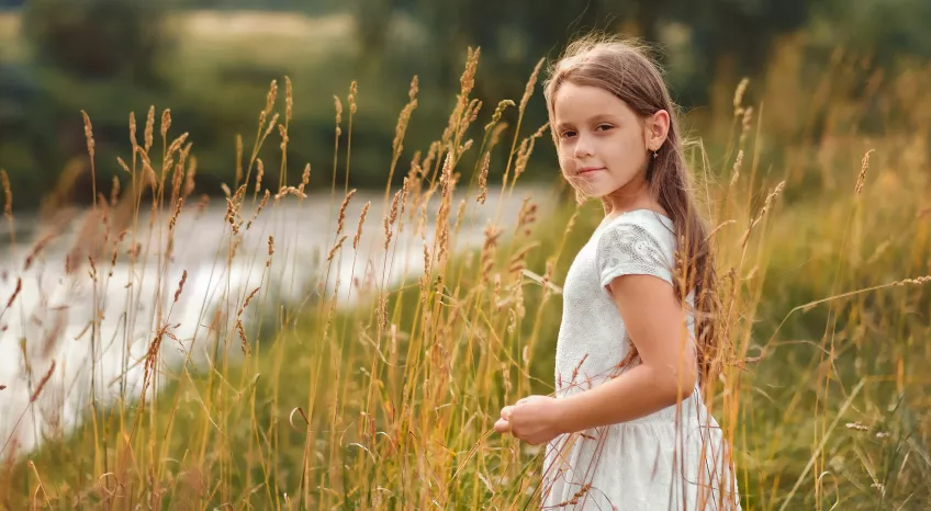 A young girl in a white dress is standing among reeds by a river.