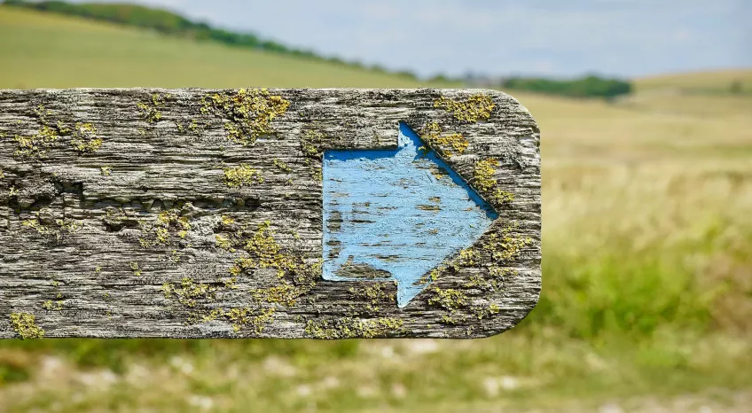 Close up of an old wooden road sign with a blue arrow pointing right painted onto it. In the background is moorlands.