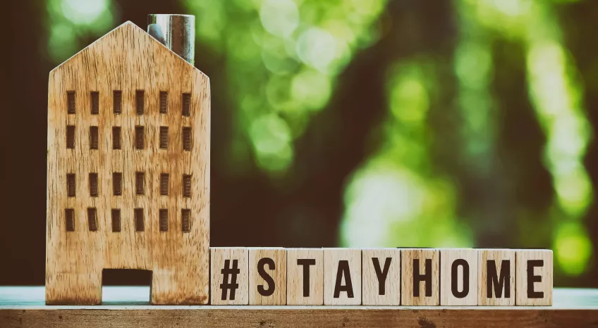 A close up of a wooden toy house and wooden letter boxes spelling out #STAY HOME. The backdrop is green leaves with bokeh efect.