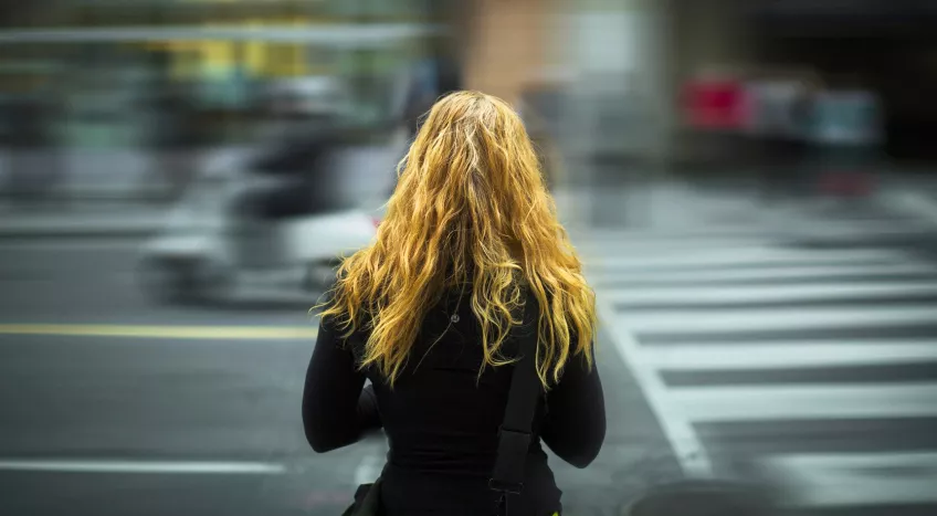 A woman with blonde hair is photographed from behind. She's standing by a zebra crossing and the traffic is rushing past in front of her