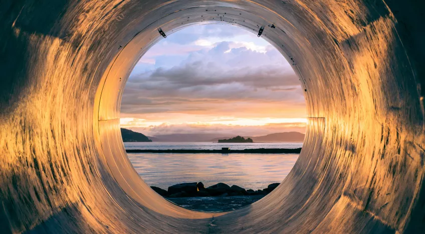 POW standing inside a large pipe, looking out on a rocky beach, the sea, and beyond: a sunset 