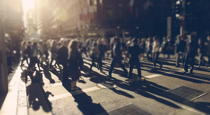 Lots of people are crossing a busy intersection. The light is soft and against the camera, so the people are blurry. 